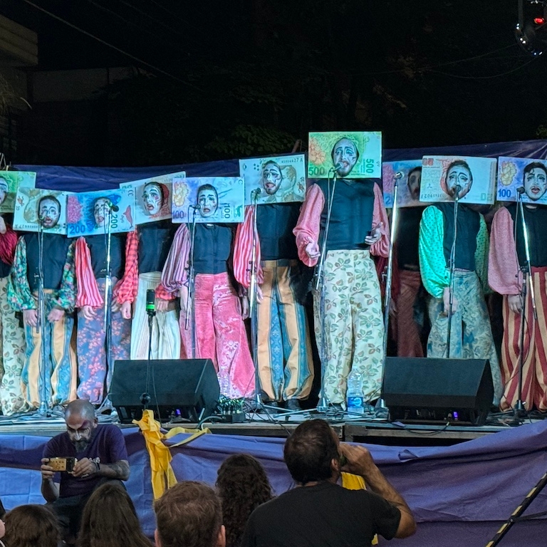 Murga performers on stage wearing vibrant costumes and currency face cutouts that show white painted faces.